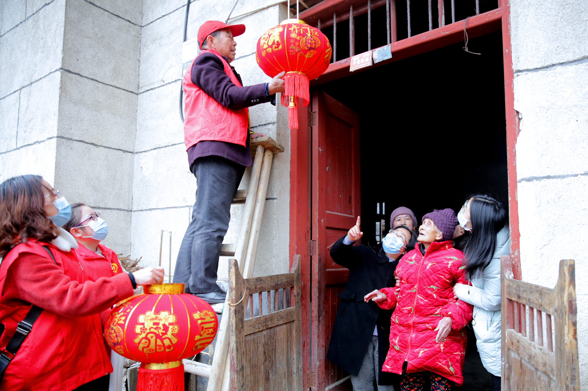 图为2月1日，桂东县沤江镇三洞村陈瑞鸾家大门口，陈瑞鸾（右二）在志愿者挽扶下看志愿者帮她挂灯笼，浓浓的年味扑面而来。  (1).JPG